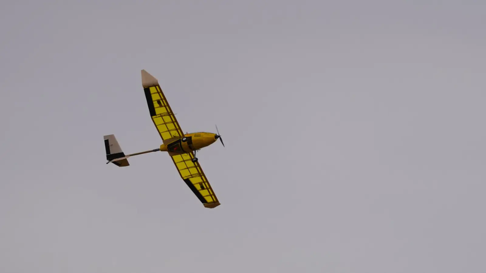 Velículo de ala fija amarillo y negro en vuelo contra un cielo cubierto, hélice en rotación, costillas visibles bajo la película.